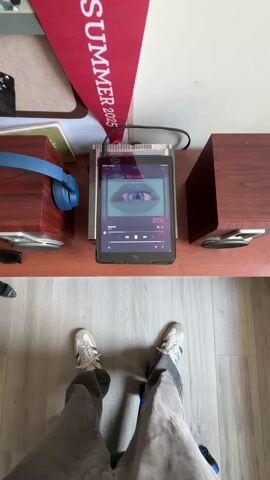 The video presents a static, high-angle shot of a person standing over a wooden shelf. The person is wearing grey pants and white and blue sneakers. On the shelf, from left to right, is a wooden speaker, blue headphones, a pair of sunglasses, several books, a black tablet connected with a cable to an amplifier, and another wooden speaker. The tablet displays music playing, with the song title "Nena" and the artist "Zoe" visible. Above the shelf, a red banner with the words "SUMMER 2005" can be seen. The floor beneath the person is light-colored wood. The scene is well-lit, but there's no apparent camera movement. The focal point is the arrangement of objects on the shelf.