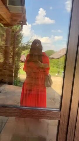 The video shows a reflection in a glass door on a sunny day. The reflection captures a woman wearing a red dress and a light-colored purse. She is holding a phone, suggesting she might be taking a selfie or recording a video. Behind her, the reflection reveals a rustic wooden building, possibly a house or cabin, with log accents and a visible rain gutter. The outdoor scene outside the glass includes lush green trees, a patch of grass, and a bright blue sky with fluffy white clouds, indicating pleasant weather and likely daytime. The overall setting appears to be a rural or semi-rural environment, possibly a vacation home or a countryside residence in Bogdanikha, Russia. There are no discernible signs or text within the visible frames of the video. The mood appears relaxed and casual, with the woman seemingly enjoying the outdoors.
