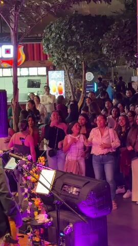 The video shows a lively outdoor gathering with a crowd of people enjoying a performance. In the foreground, a stage is set up with musical equipment including speakers and instruments, bathed in purple lighting. Several people are visible on stage, and a musician is playing a guitar. The audience, composed primarily of women, stands in front of the stage, many with their hands raised in excitement and some singing along. They are dressed in casual attire, including jeans and tops. Behind the audience, there are trees and some structures that appear to be part of a market or entertainment venue, with red shipping containers visible in the background. A sign with an image of two drinks is also present. The overall atmosphere is festive and energetic, suggesting a live music event or concert.
