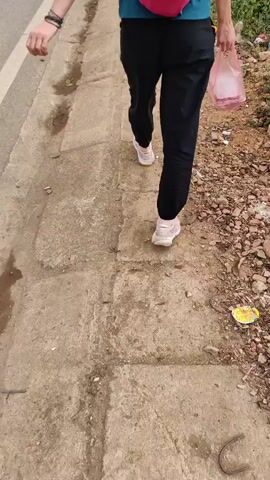 A person is walking away from the camera on a concrete sidewalk adjacent to a road. The person is wearing black pants, white sneakers, and a teal shirt, with a small pink backpack. They are carrying a clear plastic bag, possibly containing food. The sidewalk is cracked and stained, with some debris and plant growth along the edge where it meets a patch of dirt and stones. The road is visible on the left.