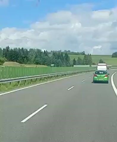 The video shows a view from a vehicle traveling on a highway. A green car with a white object on its roof is in the right lane ahead. A green sound barrier lines the left side of the highway. Trees and hills are visible beyond the sound barrier under a blue sky with white clouds. The highway curves to the right in the distance.