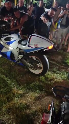 The video shows a group of men gathered around a white and blue Suzuki motorcycle. The motorcycle is parked on grass, with another motorcycle partially visible in the foreground. Several men are leaning over the Suzuki, with one man appearing to be working on the handlebars or controls. The lighting suggests it is nighttime. The men are casually dressed, and some are holding up phones, possibly recording or taking pictures. There is an energetic atmosphere, with some of the men gesturing and appearing excited.