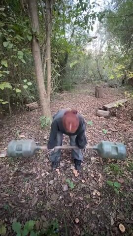 The video shows a person with red hair, wearing a gray sweatshirt and pants, standing in a wooded area covered with leaves and vegetation. The person is positioned in front of a barbell with two large green water jugs attached to each end as weights. They are in a squat position, preparing to lift the barbell. The area around them is a natural outdoor setting with trees, logs, and foliage scattered around. The lighting is slightly overcast, giving the scene a somewhat muted tone.