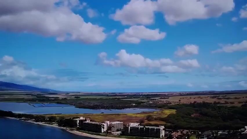 This is a drone's eye view of a tropical coastline. The ocean, in a deep blue, takes up the left third of the frame. The coastline features a narrow strip of beach leading to a cluster of multi-story buildings, possibly hotels or condominiums. Beyond the buildings, a body of calm water, perhaps a lagoon or bay, is visible, dotted with what appear to be solar panels. The background shows a vast expanse of flat land with patches of vegetation and some agricultural fields, eventually leading to distant mountains under a bright blue sky filled with fluffy white clouds. The scene appears to be in a state of partial development or perhaps some of the buildings are no longer in active use, suggested by their appearance. The overall atmosphere is one of natural beauty with signs of human infrastructure.