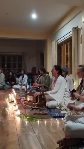 This video captures an intimate evening gathering inside a communal room, likely in Dharamkot, India. A diverse group of people, primarily young adults, are seated on a wooden floor, participating in what appears to be a kirtan or devotional music session. The room is softly lit, creating a warm ambiance, with numerous lit candles and string lights artfully arranged along the floor in the foreground.

At the center, a woman in a white dress is leading the session, singing with her head tilted back and playing a harmonium placed on a patterned rug. To her left, a man with dark, curly hair is playing a flute. To her right, another woman with braided hair is smiling and appears engaged in the singing or chanting. Several other participants are seated behind them, listening or participating. The overall atmosphere is peaceful and communal, indicative of a shared spiritual or meditative experience. The background features light-colored walls and windows with curtains, further suggesting an indoor communal space.