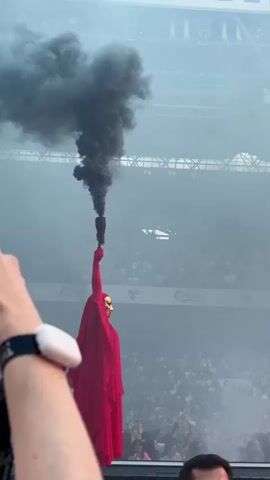 The video shows a person in a red robe and a gold mask holding a lit flare that is emitting black smoke. The person is standing in front of a crowd of people in what appears to be a stadium. The crowd is blurry and out of focus, but it is clear that there are many people present. The background is also blurry, but it appears to be a stadium setting with seating and some signage. The smoke from the flare is obscuring the upper part of the person and creating a dramatic effect. A hand with a wristband and a white object is in the foreground, on the left side of the frame, holding a phone. The video is shot from a low angle, looking up at the person with the flare.