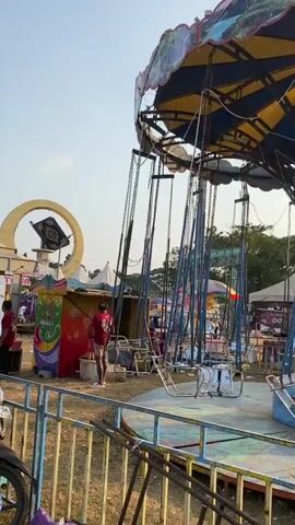 A carnival ride, likely a chair swing, is shown in the video. The ride has a central pole structure with a large, circular, yellow and blue canopy at the top. Hanging from the canopy are numerous chains attached to seats. Some of the seats are empty, while others appear to have people in them, though they are not clearly visible. The ride is situated in an outdoor setting, with a fence in the foreground and other carnival structures, tents, and trees visible in the background. The sky is clear and appears to be daytime. A person is visible standing near the base of the ride on the left side of the frame, wearing a red shirt and a mask. They are near a colorful booth or stand with graphics. The overall impression is that of a carnival or amusement park.