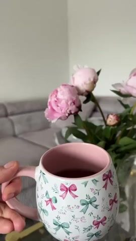 A person's hand holds a white mug with a pink rim and a pattern of pink and green bows. The mug is filled with a dark liquid. In the background, a bouquet of pink peonies in a clear glass vase sits on a glass-topped coffee table. A gray sectional sofa is visible behind the table. The scene is indoors, with a plain white wall in the background.