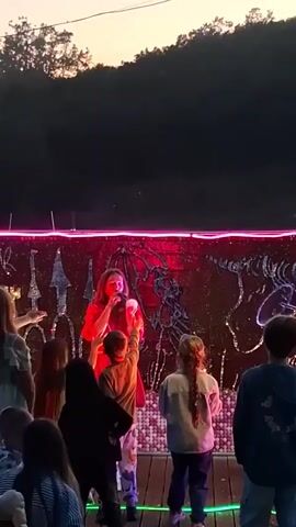A woman in a red dress is performing a bubble show for a group of children. The children are reaching for the bubbles. The performance is taking place outdoors in front of a wall decorated with skeletons and other designs, highlighted with a pink neon light.  The sun is setting in the background.