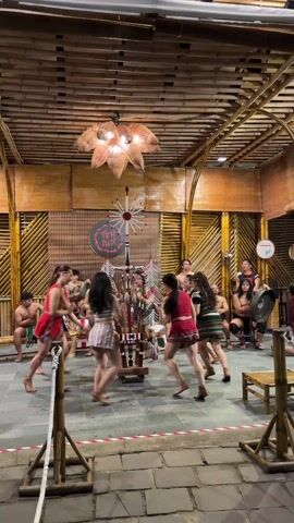The video shows a group of about ten dancers dressed in colorful, indigenous-style clothing, dancing in a circle around a decorated pole-like structure. Several men are sitting around the perimeter, some playing drums and other instruments. The room is decorated with bamboo, including the walls and ceiling, and there is a large, ornate light fixture hanging from the ceiling. A sign that says "Teh Dar" is visible in the background. The dancers are mostly women, and their movements appear rhythmic and coordinated. The atmosphere seems festive and cultural.