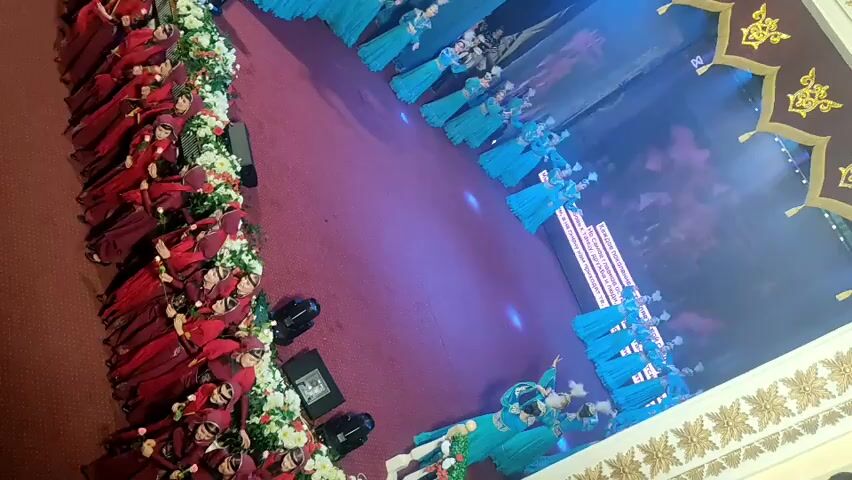 A video taken from a high angle shows a performance on a stage. On the left side of the frame, a group of people wearing red dresses and headscarves are standing in a line. In the center and right of the frame, another group of people wearing blue dresses are arranged in a semi-circle facing a stage. On the stage, there is a large screen displaying an image of people in red and blue costumes. Some people in green costumes are dancing in the foreground. The floor is a deep purple color. There are decorative elements on the right side of the frame, suggesting an ornate setting.