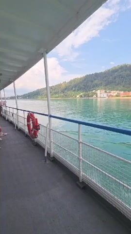 The video shows a view from a boat on a body of water. In the foreground, there is a deck with a railing. A life preserver is attached to the railing. In the background, there is a town or city along the shore, with buildings and trees. The sky is blue with some clouds. The water is a turquoise green color and appears calm.