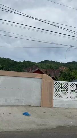 The video shows a view from a moving vehicle. The first shot is of a white, patterned fence with a hill in the background. The second shot shows the entrance to a property, with a white guardhouse and a decorative sign. The third shot is of a small, closed shop with a display case. The sky is overcast in all shots.