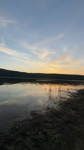 The media file captures a serene lakeside scene at sunset, likely in Debelets, Bulgaria. The sky is a gradient of soft blues transitioning to warm oranges and yellows near the horizon, streaked with wispy clouds. A dark, hilly landscape forms the backdrop, silhouetted against the vibrant sky. The lake's surface is calm, reflecting the sky and the distant hills with a mirror-like quality.

In the foreground, the muddy shore is visible, dotted with dry grass and reeds. Emerging from the water are a few tall, slender stalks of vegetation, adding a touch of natural texture. To the left of the frame, a green, collapsible fishing net rests partially submerged in the water, suggesting that fishing is either in progress or has recently occurred. A fishing rod, with its tip clearly visible and a red accent on the reel, extends from the right side of the frame, also planted near the shore, further indicating fishing activity.

The time of day is clearly sunset, given the colors in the sky and the soft, diffused light. The weather appears to be calm and pleasant, with still water indicating minimal wind. The overall emotion evoked by the scene is one of peace, tranquility, and quiet enjoyment of nature. There is no visible text within the media file.