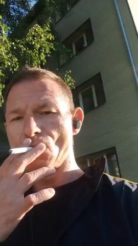 The video shows a close-up, selfie-style recording of a man smoking a cigarette outdoors. The man has short, light brown hair and is wearing a black shirt. He is holding the cigarette in his right hand and appears to be exhaling smoke. He has an earbud in his right ear. The background consists of a building with windows and some greenery, possibly trees or bushes. The lighting suggests it is daytime, with sunlight illuminating the scene.