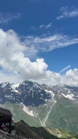 The video shows a panoramic view of a mountain landscape with snow-capped peaks and green valleys under a blue sky with scattered clouds. In the foreground, there are buildings that appear to be mountain huts or lodges with wooden structures and signs. A parking area with several cars and a van is visible, and some people are seen walking around the buildings and the parking lot. The camera pans slightly to capture the vastness of the mountains and the surrounding environment.