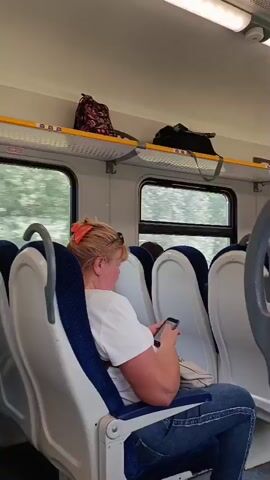 The video shows the interior of a train car. In the foreground, several rows of blue and white seats are visible. A woman with blonde hair tied back with an orange clip is seated in one of the seats, looking down at her phone. She is wearing a white t-shirt and blue jeans. Above the seats, there is a luggage rack with bags on it. The window of the train shows blurred green trees, indicating that the train is moving. There is a red emergency brake handle mounted on the wall.