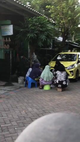 A group of four individuals, three appearing to be women and one child, are gathered outside on a paved area. Two of the women are seated on blue and green stools, while the other woman and the child are kneeling. They are facing a table, though the table is not fully visible. A yellow car is parked to the right of the scene, and lush green foliage is present behind the individuals and to the left. The lighting suggests it is daytime.