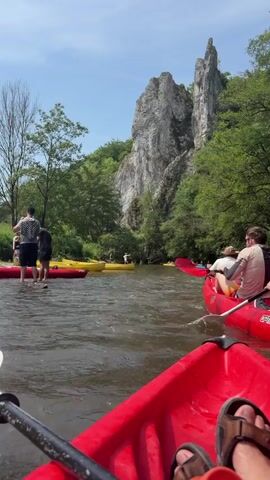 The video shows a person's point of view from a red kayak paddling down a river.  Several other kayaks, both red and yellow, are visible in the river. In the background is a tall, rocky cliff face surrounded by trees. The sky is blue with some clouds. Some people are standing in the shallow water near the kayaks. The person in the kayak is wearing sandals and using a paddle.  Two other people are in the kayak in front of the camera.  The river is moderately flowing.