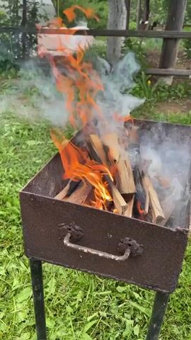 A vertical shot in a natural outdoor setting shows a fire burning in a dark metal grill with four legs. The grill is filled with wood pieces, some of which are consumed by flames. The fire is mainly concentrated at the top and middle of the wood, with orange flames reaching high and some smoke rising on the right side. The grill is placed on green grass, and in the background, a wooden fence and various plants are visible. The sun is shining, and the overall scene depicts a peaceful backyard environment.
