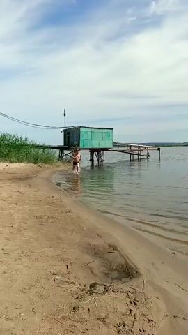 The video shows a man running into a body of water on a sandy beach. He is shirtless and wearing dark swim trunks. The water is calm and reflects the sky. In the background, there is a wooden structure on stilts over the water, which appears to be a fishing hut or a small pier. Tall reeds grow along the edge of the water to the left of the man. The sky is blue with scattered white clouds, suggesting a fair-weather day. The sun is casting a shadow from the man onto the sand, indicating it is likely daytime, possibly late morning or early afternoon. The overall scene evokes a sense of leisure and outdoor activity, likely during warmer months. The location is identified as Vyazovka, Russia. There is no visible text in the video.