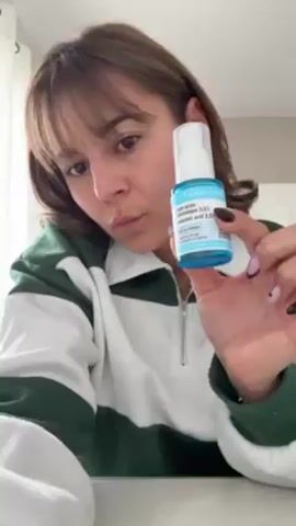 Here's a detailed description of the video:

A young woman with light brown hair and bangs is holding up a small, blue-and-white bottle of serum. The bottle is labeled "AROMA-ZONE Sérum acide hyaluronique 3.5%" and "MADE IN FRANCE." She is wearing a white sweatshirt with green stripes and gold hoop earrings. She is sitting at a table, with part of a white blind visible in the background. The woman is looking at the camera, but the focus is on the product she is holding. Her fingernails are painted light purple and dark gray.