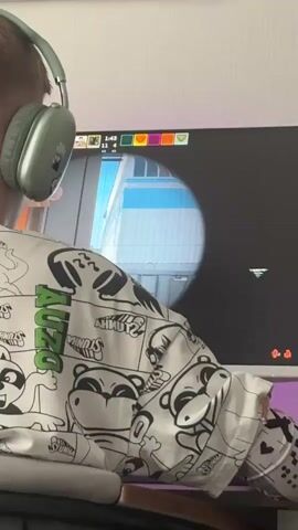 A person wearing headphones and a white long-sleeved shirt with cartoon graphics is sitting at a desk and playing a video game on a computer. The game appears to be a first-person shooter, and the player is looking through a scope. The room has a pink light behind the monitor and a picture on the wall.