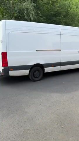 A white cargo van is parked on an asphalt surface next to a green fence and trees. The van is facing away from the camera. The rear doors of the van are closed.