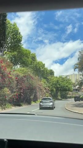 The video shows a car driving on a road. The view is from the inside of the car, looking out the windshield. There is a silver car in front. The sky is blue with white clouds. There are trees and buildings on the side of the road.