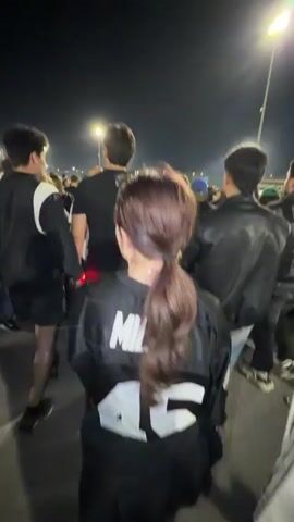 At night, a large crowd of people walks away from an event. Streetlights provide illumination. Most of the crowd members wear dark clothing. The camera follows behind a woman with reddish hair pulled into a ponytail. She wears a black jacket with white lettering on the back. Two people in black jackets walk on either side of her. Other crowd members are also visible walking and moving away from the camera's perspective. Trees are visible on the left and smoke is discernible in the background.