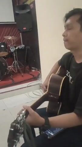 A medium shot captures a group of people in what appears to be a church or community hall. The scene is well-lit, with a bright, possibly tiled floor. In the foreground, on the left, a person is playing an acoustic guitar. To their right, a smartphone rests on their lap. Behind them, a drum set is visible, set up on a red carpet. Further back, a woman sits on a chair, facing the camera, with her hands clasped in her lap. To her right, a young woman is also seated, with her legs crossed and feet bare. To the far right, another woman is visible, also seated. The background features a red wall adorned with religious imagery, including a large cross with flowers. The overall atmosphere suggests a gathering or a rehearsal for a musical performance.