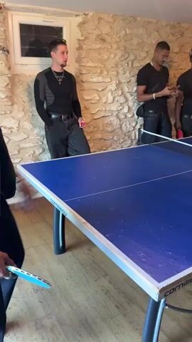 Four Black men are gathered around a blue and white ping pong table in an indoor setting. Three of them are standing while one plays a shot. The walls are of a beige stone and there is an unopened can of table tennis balls near the player. The men are all casually dressed. One is looking at his phone. The lighting in the room is dim and there is wood flooring.