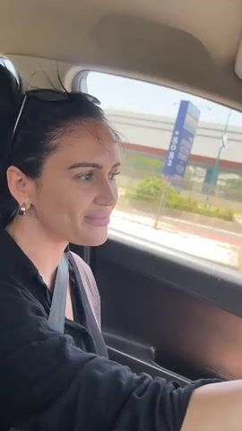 The video shows a person driving in a car, focusing on the interior and the view through the windshield.

The driver is a woman with dark hair pulled back, wearing a black shirt and a seatbelt. She is driving and looking to her left, then forward. Sunglasses are perched on her head. She has a silver earring and a wedding ring on her left hand. 

The car's interior is visible, including the steering wheel, dashboard, rearview mirror, and the passenger side. A small decorative item hangs from the rearview mirror.

The exterior view through the windshield shows a sunny day with palm trees lining the road. Buildings, other cars, and street lights are also visible. The video appears to be taken in a commercial area.