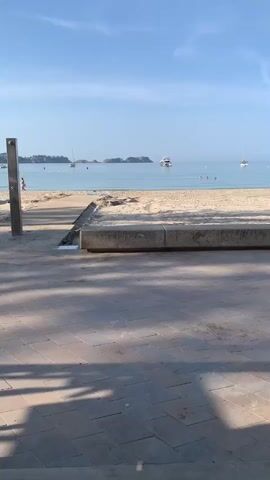The video shows a wide shot of a beach with a paved walkway in the foreground. The sky is clear blue with a few clouds. The ocean is calm with several boats and buoys visible in the distance. The beach is sandy and appears to be mostly empty. There is a concrete bench or wall in the middle ground and a metal shower fixture on the left side of the frame. Shadows from the walkway are cast across the paved area. The overall impression is a peaceful, sunny day at the beach.