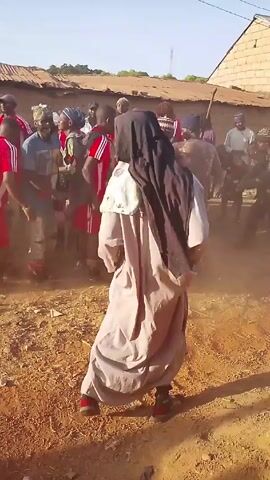 The video shows a group of people gathered in a dusty, outdoor area. Most of the people are wearing casual clothes, but one person is wearing a long, grey robe and a black head covering. This person is walking toward the group of people, who are dancing and celebrating. The overall atmosphere seems festive. The video is shot from a low angle, which makes the people seem larger and more imposing. In the background, there are buildings with corrugated iron roofs.