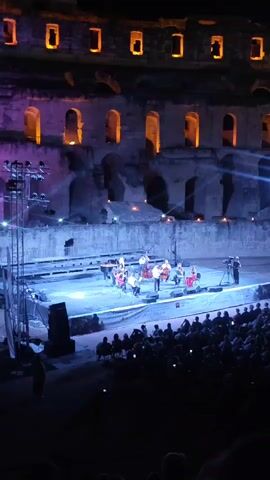 The video shows a performance at night in an amphitheater. A group of musicians and possibly dancers are on a brightly lit stage, with a piano, drums, and other instruments visible. The amphitheater's ancient stone structure is illuminated with purple and orange lights, highlighting arched openings. A crowd of spectators is seated in the foreground, facing the stage, and some are holding up their phones to record the event. The overall atmosphere appears to be a cultural or musical performance in a historic setting.