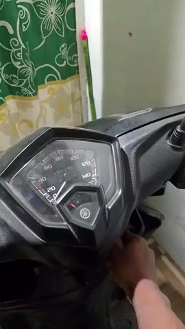 A person's hand is seen turning the ignition key of a black Yamaha motorcycle. The motorcycle's dashboard lights up, displaying the speedometer, odometer, and fuel gauge. The speedometer needle moves briefly as the ignition is turned. The background consists of a green and white floral curtain and a wall.