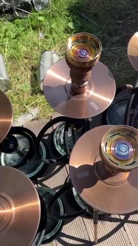 The video shows a top-down view of several hookahs arranged on a wooden deck outdoors. The hookahs have copper-colored bases and ornate brown stems. Their bowls contain colorful, multi-layered inserts, and one bowl has white charcoal cubes being placed into it by a person wearing black gloves and holding tongs. The environment includes green grass and a blurred background that appears to be an outdoor seating area. The camera pans slightly and zooms in at different points to highlight the hookahs and the charcoal placement.