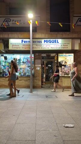 A woman in a long grey dress walks along a sidewalk at night. She is to the right of the frame, moving away from the camera. In the background, a store called "Ferreterias MIGUEL" is visible with products displayed in the window. Several other people are walking in the same direction as the woman in the grey dress. The sidewalk is made of concrete slabs, and there is a street lamp overhead. The scene is dimly lit by the street lamp and the lights from the store.