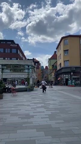 The video captures a wide shot of a bustling pedestrian street in Uppsala, Sweden, on a clear day with a bright blue sky and prominent white, fluffy clouds. The street is paved with light-colored, rectangular tiles arranged in a decorative pattern.

On the left side of the street, a modern, multi-story building with a brown/red brick facade and large windows is visible, featuring the "Ductus" logo. Below it, a building houses "Bröd & Salt," likely a cafe or bakery, with an outdoor seating area. Several people are gathered here, seated at tables under a canopy adorned with greenery, enjoying the outdoor setting.

Across the street, on the right, an older, multi-story building with a yellow facade stands. Further down the street, a cluster of buildings with diverse, colorful facades and unique architectural details, including what appear to be decorative turrets or roof elements in shades of green and red, adds to the charm of the scene.

Several individuals are walking along the pedestrian thoroughfare; one person is prominently walking towards the camera from the middle of the street. Others are seen strolling in the distance or seated at the outdoor cafe. The overall ambiance suggests a lively, pleasant urban environment during daytime. Two green bins or kiosks are also visible on the far right side of the street.