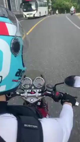 A motorcyclist wearing a light blue and white helmet and a Burton backpack rides on a gray asphalt road. The camera is positioned behind the rider, capturing a view over their shoulder. The rider's hands, clad in black gloves, are visible on the motorcycle's handlebars. The motorcycle has a dark red gas tank and an instrument panel with two gauges. Other vehicles, including a white bus and other motorcycles, are seen on the road ahead. The road curves slightly and is marked with a solid white line. On the right side of the road, green vegetation is visible. A sign with blue lettering is mounted on a metal pole on the left side of the road. As the rider continues, a sign in Vietnamese becomes visible. The sky above is overcast with gray clouds.