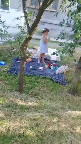 This media file captures a young child playing outdoors. The child, dressed in a light blue dress, is standing on a dark mat surrounded by various toys. These include what appear to be dolls, colorful balls, and a blue plastic container. A doll's head is visible on the mat. The ground is covered with grass and dry leaves, suggesting it might be late spring, summer, or early autumn. Behind the child and the play area, there's a building with windows and a grey facade. The scene is lit by natural light, indicating it's daytime, possibly afternoon given the lighting and shadows. The child's posture suggests engagement with the toys, potentially engrossed in play. The overall setting appears to be a residential outdoor area, perhaps a courtyard or a backyard, with some vegetation and a tree in the foreground partially obscuring the view. The location context is Novyi Rozdil, Ukraine. No visible text is present in the media.