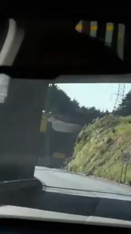 A car is driving down a highway on a sunny day. The driver is looking at a sign that reads "RAMPA DE EMERGENCIA 1 A 1 Km," which translates to "EMERGENCY RAMP 1 AT 1 Km" in English. The sign is located above a road that is curving into a concrete tunnel, and there are power lines and trees visible in the background.