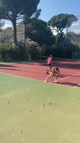 Two young females are playing tennis on an outdoor court. The court has a red surface with green out-of-bounds areas. The players are wearing athletic clothing. One player in a purple top and black shorts is in the foreground, running and swinging a tennis racket. The other player in a pink top and black shorts is on the opposite side of the court, also in motion. A third person, mostly obscured, is visible in the background on the same side as the player in pink. The scene is set under a clear blue sky with trees and a tennis net in the background. There are also shadows cast on the court, indicating it is daytime. A ladder and a bench are also visible on the edge of the court.