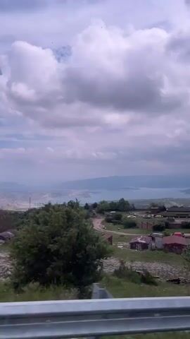 The video shows a landscape from a moving vehicle, with rolling green hills, scattered buildings, and a large body of water in the distance. The sky is filled with puffy white clouds, and there are trees interspersed throughout the landscape. Power lines are visible in the background.