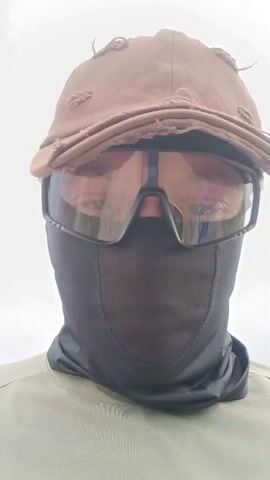 A person is facing the camera outdoors. They are wearing a brown baseball cap, dark goggles, and a black face mask that covers their nose and mouth. The person is also wearing a light brown shirt. The background appears to be a landscape with hills and sky. The video is a close-up shot of the person's face and upper body.