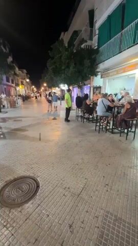 This is a shaky, handheld video taken at night on a street lined with shops and outdoor cafes.  The camera is moving and panning, creating a blurred effect. Several people are seated at tables outside the cafes, appearing to be dining or socializing. Some individuals are walking along the street. Streetlights and lights from the shops illuminate the scene. The ground appears to be paved, and there are some wet patches, possibly from recent rain. A man in a red shirt and shorts walks towards the camera in the foreground. A man in a bright green shirt is visible further down the street. The overall atmosphere seems to be one of a lively evening in a commercial area.