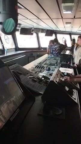 The video shows the interior of a ship's bridge. A group of people are standing around a large control panel, which is filled with buttons, levers, and screens. Two people are actively engaged with the controls, appearing to steer the ship. The bridge has large windows that offer a view of the outside, though it is unclear what is visible. The overall atmosphere seems to be one of observation and operation of the vessel.
