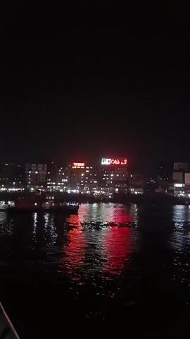 The video depicts a nighttime scene featuring a body of water reflecting the lights of a city skyline. Two prominent signs, one red and one green reading “CABLES”, are illuminated against the dark buildings. Several boats can be seen moving on the water, their lights also reflected in the ripples. The overall ambiance is dark, with the city lights and reflections providing the only significant illumination.