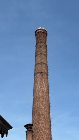 A tall brick chimney stands against a clear blue sky. The chimney is made of reddish-brown bricks and has a series of metal rungs running up its side, likely a ladder. At the top of the chimney, there is a decorative cap. In the lower left corner of the frame, a portion of a wooden structure with decorative eaves is visible. A shorter, similar chimney with a cap is also partially visible below and to the left of the main chimney. Several small dark specks, possibly birds, are scattered in the sky. The video appears to be a static shot, with no apparent movement or action.